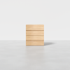 A minimalist, light wood chest of four drawers stands alone on a smooth, gray floor against a plain white background.