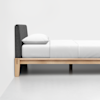 A minimalist bed with a light wooden frame, a dark upholstered headboard, and white bedding with a pillow, positioned against a plain white wall and floor.