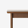 A close-up view of a wooden bench with a light-colored cushioned seat, shown against a plain white background. Only part of the bench is visible, focusing on the edge and legs.