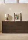 A modern wooden dresser with six drawers is topped with a ceramic lamp, a decorative tray, a small cup, and a framed abstract artwork. The dresser sits against a beige wall beneath high windows.