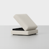 A modern, light beige ottoman with a hinged lid partially open, revealing hidden storage space inside, set against a plain white background.