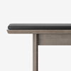 A close-up of a wooden bench with a rectangular, light wood frame and a dark gray cushion on top, set against a plain white background.