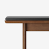 A close-up of a wooden bench with a dark gray cushion on top, set against a plain white background. The bench has clean, modern lines and a smooth finish.