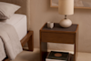 A wooden nightstand beside a neatly made bed holds a ceramic cup and a round white lamp. Books, including one titled The Japanese Garden, are stacked on the lower shelf. The setting is calm and minimalist.
