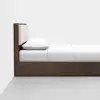 A modern wooden bed frame with a padded headboard, white bedsheet, and a pillow, shown from the side against a plain white wall and grey floor.