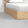 A close-up view of a light wood bed frame with a white mattress, white sheets, and a white pillow, set on a smooth light gray floor.