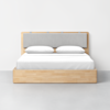 A modern wooden bed frame with a light wood finish, featuring a gray upholstered headboard, neatly made with white bedding and two white pillows, set against a plain white wall and gray floor.