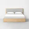 A modern wooden bed frame with a light wood finish, featuring a gray upholstered headboard, neatly made with white bedding and two white pillows, set against a plain white wall and gray floor.
