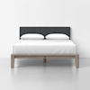 A modern bed with a wooden frame, white bedding, two pillows, and a dark upholstered headboard is placed against a plain white wall on a light-colored floor.