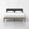 A modern bed with a wooden frame, white bedding, two pillows, and a dark upholstered headboard is placed against a plain white wall on a light-colored floor.