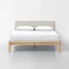 A modern wooden bed frame with a beige upholstered headboard, neatly made with white bedding and two white pillows, is set against a plain white wall and floor.