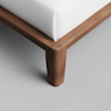 Close-up of a wooden bed frame corner with a white mattress, featuring smooth, rounded edges and precise joinery on the light-colored wood leg, set on a light gray floor.