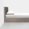 A minimalist bed with a wooden frame, a dark upholstered headboard, white bedding, and a single white pillow, positioned against a white wall on a light gray floor.