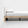 A minimalist bed with a wooden frame, white bedding, and a single pillow, positioned against a plain white wall on a smooth, light-colored floor.