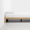 A minimalist bed with a light wooden frame, white bedding, and a single white pillow, positioned against a plain white wall in a clean, modern room with a smooth, gray floor.