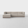 A modern L-shaped beige sectional sofa with low backrests and armrests, set on a wooden base, placed in a minimal, light-colored room with a plain background.