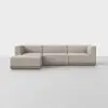 A modern L-shaped beige sectional sofa with low backrests and armrests, set on a wooden base, placed in a minimal, light-colored room with a plain background.