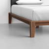 A close-up of the corner of a wooden bed frame with clean, modern lines, white bedding, and a gray headboard, placed on a light gray floor against a white wall.