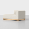 A modern, minimalist beige chaise lounge with a low wooden base and an L-shaped backrest, set against a plain white background.