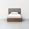 A modern wooden bed with a simple frame and a padded gray headboard, dressed with white bedding and a single pillow, stands against a plain white wall on a light gray floor.