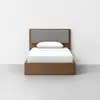 A modern wooden bed with a simple frame and a padded gray headboard, dressed with white bedding and a single pillow, stands against a plain white wall on a light gray floor.