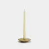 A lit cream-colored taper candle stands in the center of a round, flat, brass candle holder against a plain white background.