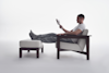 A person in a white t-shirt and gray sweatpants relaxes in a cushioned chair with their feet up on an ottoman, reading a book in a minimal, light-filled setting.