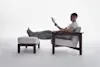 A person in a white t-shirt and gray sweatpants relaxes in a cushioned chair with their feet up on an ottoman, reading a book in a minimal, light-filled setting.