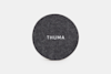 A round, dark gray felt pad with the word THUMA in white capital letters centered on it, displayed against a plain white background.