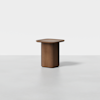 A small, modern wooden side table with a rounded square top and a solid cylindrical base, placed on a plain light gray surface with a white background.