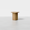 A small, modern wooden side table with a square top and rounded edges, featuring a solid, rectangular base. The table is set against a minimal white background.