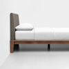 A minimalistic wooden bed frame with a gray upholstered headboard, neatly made with white bedding and a single white pillow, placed against a plain white wall.