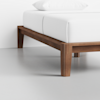 A close-up of a modern wooden bed frame with clean lines and a white mattress and pillow, set in a minimal, bright room with a light gray floor and white walls.