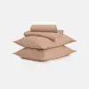 Two beige pillows stacked with a folded beige bedsheet and pillowcase set placed on top, all set against a plain white background.