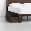 A dark wood bed frame with a white mattress and pillows, featuring an open storage drawer built into the side of the frame, set against a light-colored wall and floor.