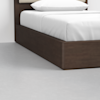 A close-up of a modern bed with a dark wooden frame, white bedding, and a white pillow, set on a light gray floor against a plain white wall.