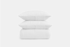 Two white pillows stacked on top of a folded white blanket, all with a subtle textured pattern, against a plain white background.