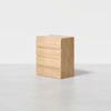 A light wood chest of four drawers stands alone on a smooth, light-colored floor against a plain white background. The design is minimalist with clean lines and no visible handles.
