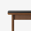 A close-up of a wooden bench with straight legs and a dark gray cushion on top, set against a plain white background.