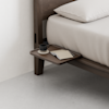 A minimalist bedside table attached to a wooden bed frame holds a closed notebook, pen, and a black ceramic cup and saucer. The bed is made with light-colored bedding, and the room has a clean, modern look.