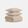 Two beige, textured pillows are stacked on top of a matching folded blanket, all set against a plain white background.