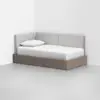 A wooden daybed with a low frame, white bedding, and a single pillow, featuring light grey upholstered back and side panels, placed in the corner of a minimalist room with white walls and floor.