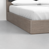 A close-up view of the corner of a wooden platform bed with crisp white bedding and pillows, set against a smooth light gray floor and a plain white wall.