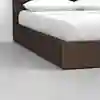 A close-up view of the corner of a wooden bed frame with a brown finish, featuring a white mattress and pillows, set on a smooth, light gray floor against a white wall.