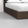 A close-up view of the corner of a wooden bed frame with a brown finish, featuring a white mattress and pillows, set on a smooth, light gray floor against a white wall.