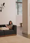 A woman sits on a dark gray sofa with a blanket, reading a book in a modern, minimalistic living room with large windows and neutral decor.