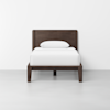 A dark wooden twin bed frame with a matching headboard, made up with white bedding and a single white pillow, is placed against a plain white wall in a minimalistic room.