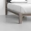A close-up view of a wooden bed frame with light brown legs and a neatly made bed with white bedding, set on a smooth gray floor in a minimalistic room.