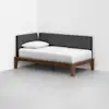 A minimalist daybed with a wooden frame, white mattress, single white pillow, and dark gray upholstered backrest, placed in the corner of a white room with a light gray floor.