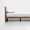 A minimalist wooden bed frame with a white mattress, white bedsheet, and a white pillow, positioned against a plain white wall and floor.